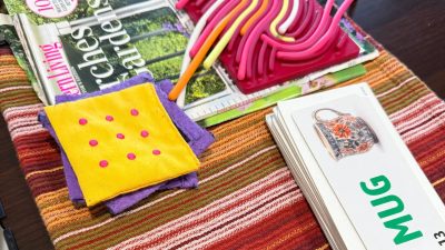 A colorful tabletop used for caregiving, featuring a Southern Living magazine, a deck of reading flashcards with MUG visible on top, a pink and yellow sensory push-and-peel toy, and small fabric squares.